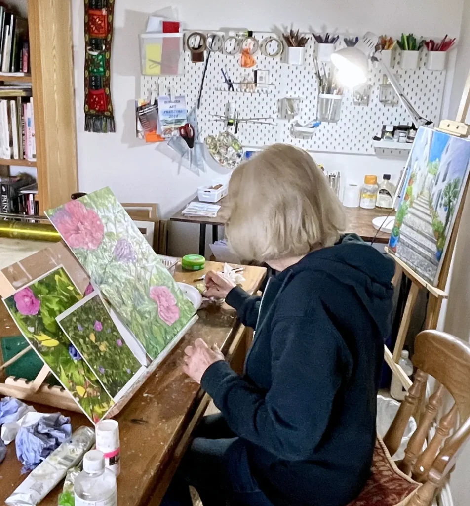 Joan Hudson, East Yorkshire artist, painting at her studio desk surrounded by art supplies with a colourful floral work in progress on the easel