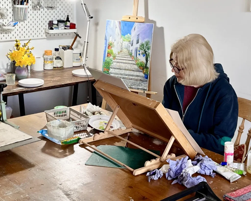 Joan Hudson, East Yorkshire artist, at work in her studio with a Tenerife street scene painting on the easel and spring daffodils on the windowsill nearby