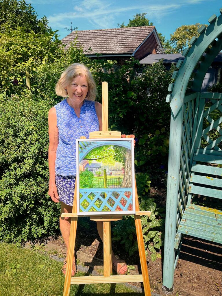 Joan Hudson standing in her summer garden beside an easel holding a garden scene painting in progress, with blue sky and greenery behind