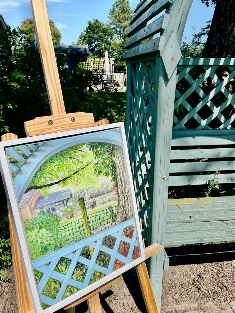 Joan Hudson's garden scene oil painting displayed on a wooden easel beside a teal painted garden seat in a sunny summer garden